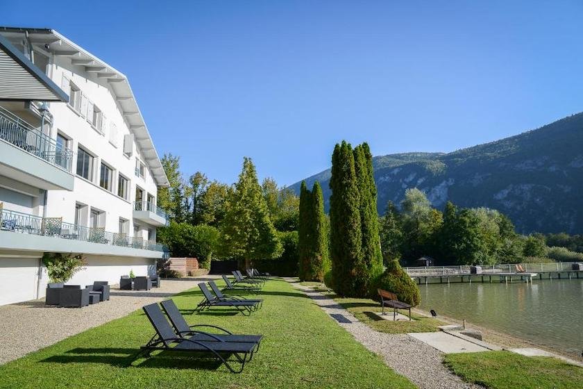 La façade blanche de l’Hôtel Novalaise Plage avec sa plage enherbée et ses chaises longues au pied du lac